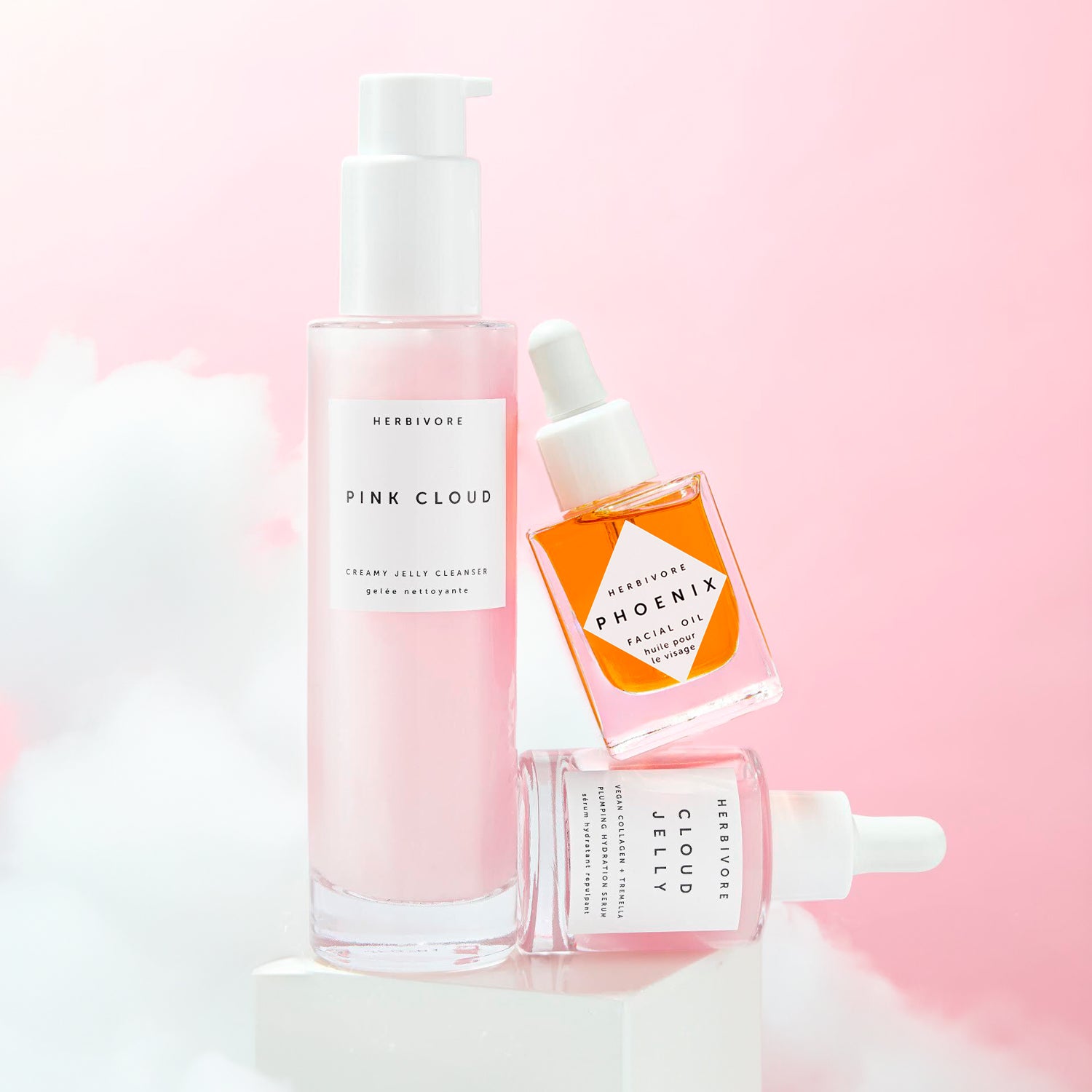 Pink Cloud Cleanser next to a stacked mid-size Phoenix Face Oil on a sideways Cloud Jelly Serum with white clouds and pink background