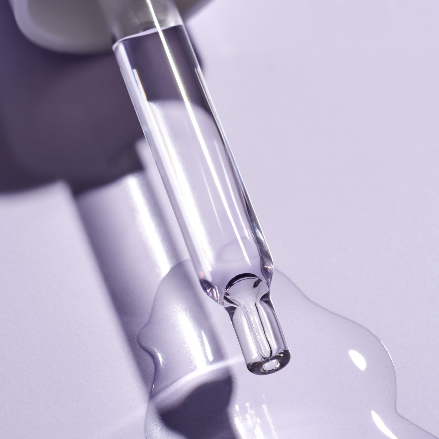 Close-up of glass dropper filled with light purple jelly-like Bakuchiol Retinol Alternative Smoothing Serum and drops of serum on a purple surface